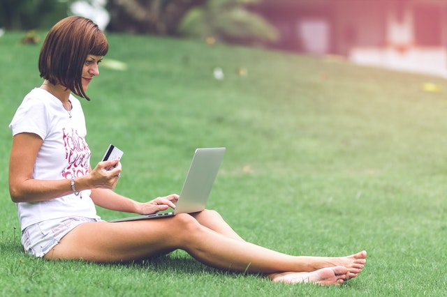 Žena s bankomatovou kartou v ruke sedí na tráve a pozerá do notebooku